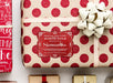 A red glitter cardstock Santa gift tag is shown on a Christmas present. The present paper is made of craft paper and has glittery red polka dots. A gold bow and ribbon are around the present. The box is seen on a white surface with other presents.