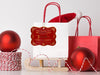 A gold foil cardstock Santa gift tag is shown hanging on a white gift bag. The bag is sitting on a sled and is surrounded by Christmas ornaments, a Santa hat, and a red gift bag. Everything is set against a white backdrop.