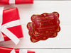 Two gold foil cardstock Santa gift tags are shown on a white wooden surface. Three red and white wrapped gift boxes are shown around the tags.