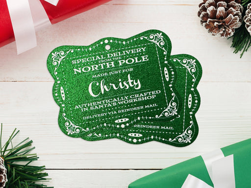 Two green glitter cardstock Santa gift tags are shown on a white wooden surface. Pine tree branches, pine cones, and Christmas presents are shown around the tags.