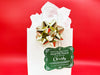 A green glitter cardstock Santa gift tag is shown hanging on a white gift bag. The bag is on a red backdrop and has a gold bow on it.