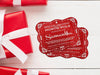 Two red glitter cardstock Santa gift tags are shown on a white wooden surface. Three red and white wrapped gift boxes are shown around the tags.