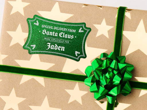 A green glitter cardstock Santa gift tag is shown on a Christmas present. The present paper is made of craft paper and has gold stars on it. The box is seen on a white surface.