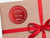 A red glitter cardstock Santa gift tag is shown on a craft paper wrapped box. The box also has a red ribbon wrapped around it. The box sits on a white wooden surface.