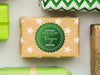 A green glitter cardstock Santa gift tag is shown on a Christmas present. The present paper is made of craft paper and has white stars on it. The box is seen on a white surface with other presents around it.