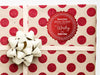 A red glitter cardstock Santa gift tag is shown on a Christmas present. The present paper is made of craft paper and has glittery red polka dots. A gold bow and ribbon are around the present. The box is seen on a white surface.