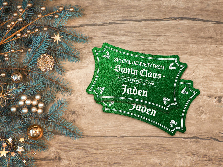 Two green glitter cardstock Santa gift tags are shown on a wooden table. The table has gold Christmas ornaments and pine tree branches on it.