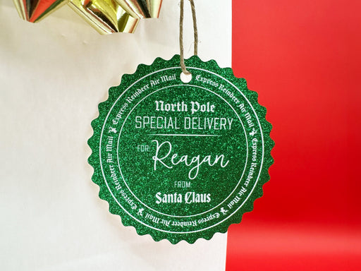 A green glitter cardstock Santa gift tag is shown hanging on a white gift bag. The bag is on a red backdrop and has a gold bow on it.