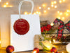 A gold foil cardstock Santa gift tag is shown hanging on a white gift bag. The bag is sitting on a wooden table, surrounded by lights, Christmas ornaments, and other Christmas elements.