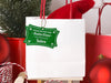 A green glitter cardstock Santa gift tag is shown hanging on a white gift bag. The bag is sitting on a sled and is surrounded by Christmas ornaments, a Christmas tree, and a red gift bag. Everything is set against a white backdrop.