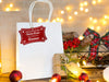 A red glitter cardstock Santa gift tag is shown hanging on a white gift bag. The bag is sitting on a wooden table, surrounded by lights, Christmas ornaments, and other Christmas elements.