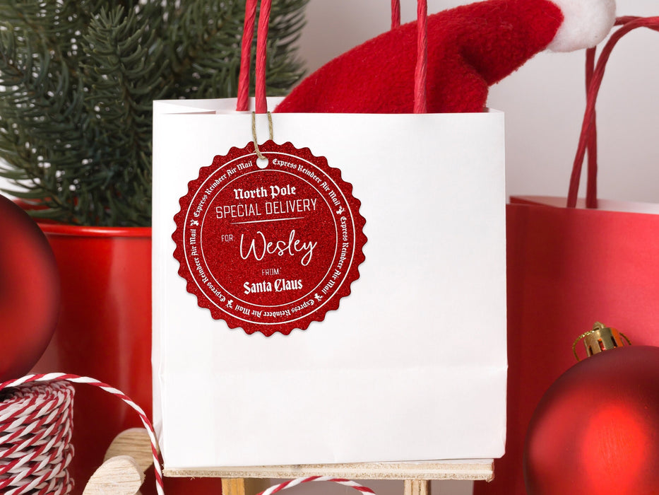 A red glitter cardstock Santa gift tag is shown hanging on a white gift bag. The bag is sitting on a sled and is surrounded by Christmas ornaments, a Christmas tree, and a red gift bag. Everything is set against a white backdrop.