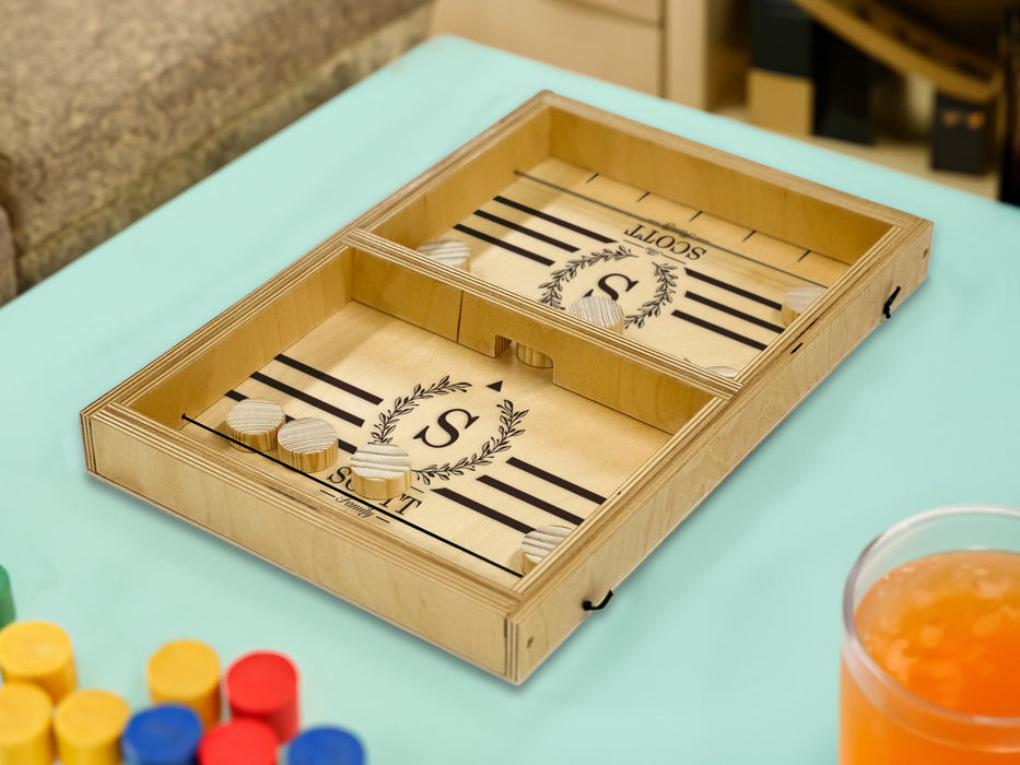 puck sling board game with an S monogram with wreath design that reads The Scott Family on light blue table surrounded by an orange drink and multicolored game pieces