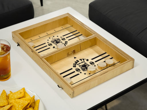 puck sling board game with wolf animal mascot that reads Silas Tys Puck Sling 2009 on white table surrounded by a glass cup of tea and a bowl of chips as well as black cushioned couches
