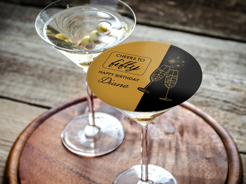 One coaster is shown being used as drink cover on top of a martini glass. Coasters are designed in black and gold ink with cheersing champagne glasses. Coaster text reads Cheers to Fifty. Happy Birthday Diane.