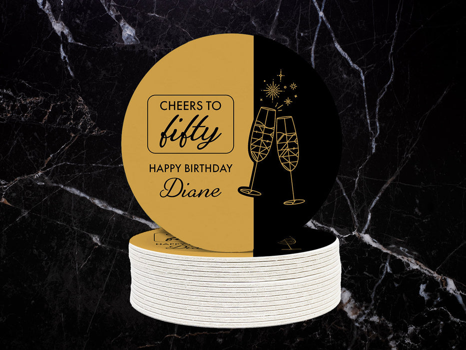 A stack of coasters are shown against a black and white marble surface. Coasters are designed in black and gold ink with cheersing champagne glasses. Coaster text reads Cheers to Fifty. Happy Birthday Diane.