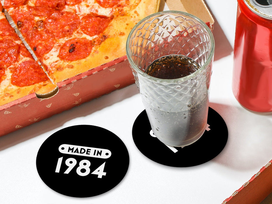 One coaster has a drink on it and an empty coaster sits beside it on a white table with pizza boxes. Coasters are designed with a custom year. Coasters read MADE IN 1984 and designed with white text and black background.