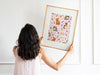 woman with black hair and pink shirt holding up a wooden frame with an easter print of pastel bunny to hang onto wall