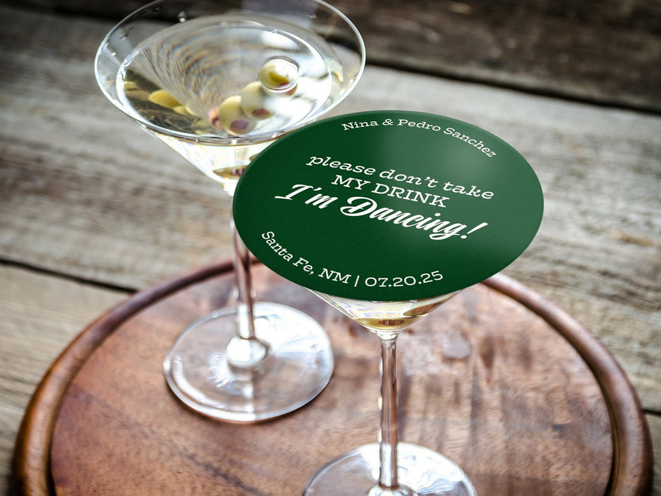 One coaster is being used as a drink cover on top of a martini glass on a wooden tray and table. Coaster says Please don't take my drink, I'm dancing with wedding couple's names, location, and wedding date.