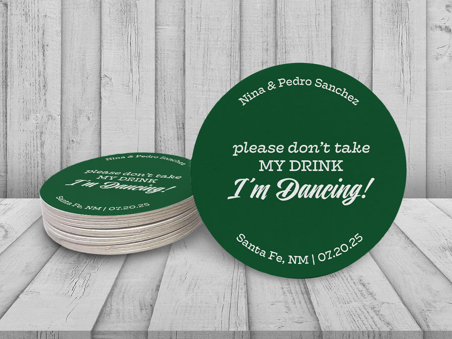 A single coasters beside a stack of coasters are shown against a white wooden background. Coasters say Please don't take my drink, I'm dancing with wedding couple's names, location, and wedding date.