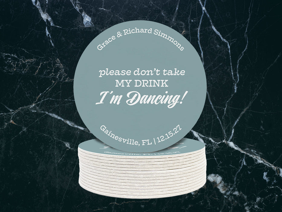 A single coasters sits on top of a stack of coasters against a dark marble background. Coasters say Please don't take my drink, I'm dancing with wedding couple's names, location, and wedding date.