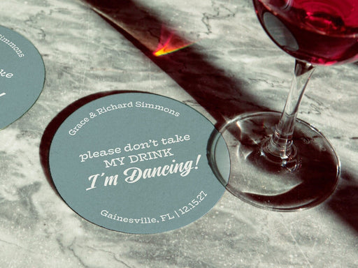 Coaster is shown with a wine glass on top of it and another off to the side. Coasters say Please don't take my drink, I'm dancing with wedding couple's names, location, and wedding date.