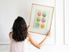 woman with black hair and pink shirt holding up a wooden frame with an  pastel easter art print of decorated eggs with a baby chick