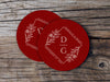A couple of red coasters sitting on top of a wooden table. Coasters feature a floral diamond design with a monogram, wedding couple's names, and wedding date.