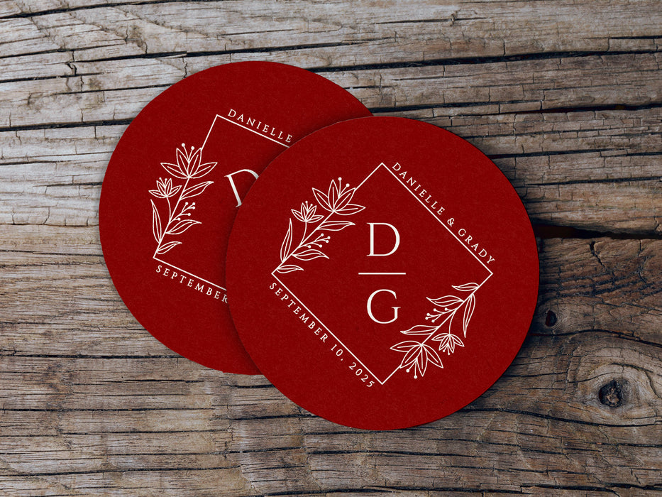 A couple of red coasters sitting on top of a wooden table. Coasters feature a floral diamond design with a monogram, wedding couple's names, and wedding date.