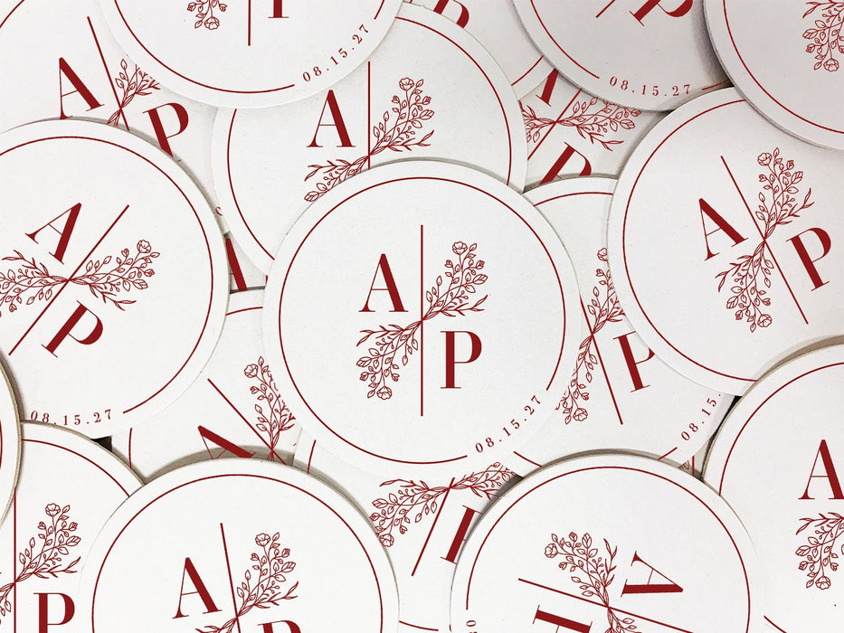 A pile of custom round coasters is shown. Coasters feature a custom floral monogram design with two initials and floral decoration in the middle and a border with a wedding date on the side.