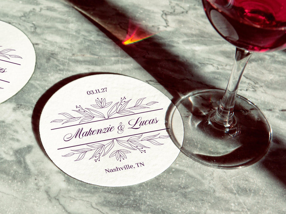 Coaster is shown with a wine glass on top of it and another off to the side. Coasters feature a personalized floral design with the happy couple's first names, wedding date, and location.