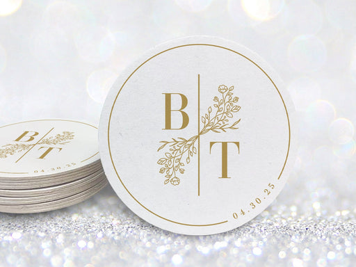 A stack of coasters by a single coaster in front of glittery background. Coasters feature a custom floral monogram design with two initials and floral decoration in the middle and a border with a wedding date on the side.