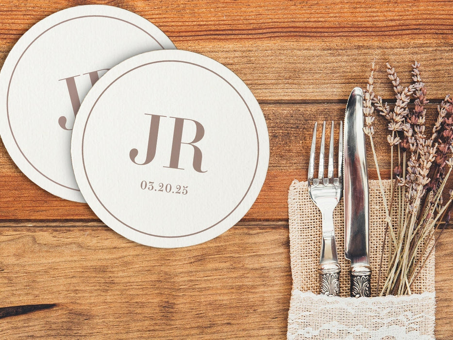 A couple of custom coasters sitting on top of a wooden table next to silverware. Coasters feature a personalized monogram design with a couple's first name initials and date.