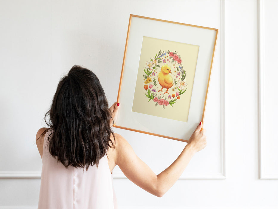 woman with black hair and pink shirt holding up a wooden frame with an  pastel easter art print of a baby chick surrounded by spring flowers