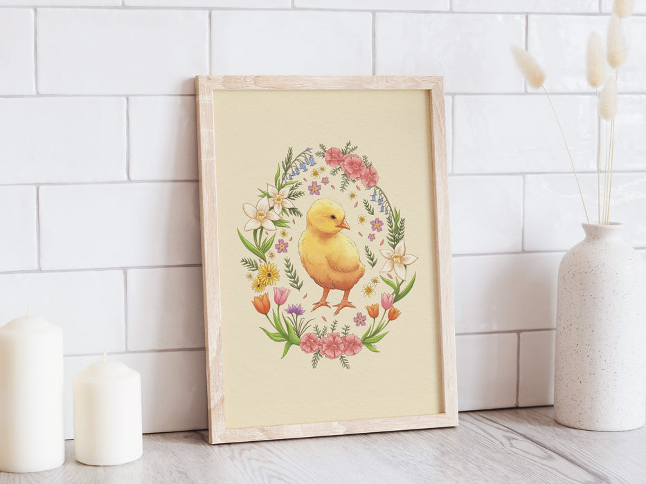 wooden frame with pastel easter art print of a baby chick surrounded by spring flowers on a white kitchen counter top surrounded by candles and a potted plant
