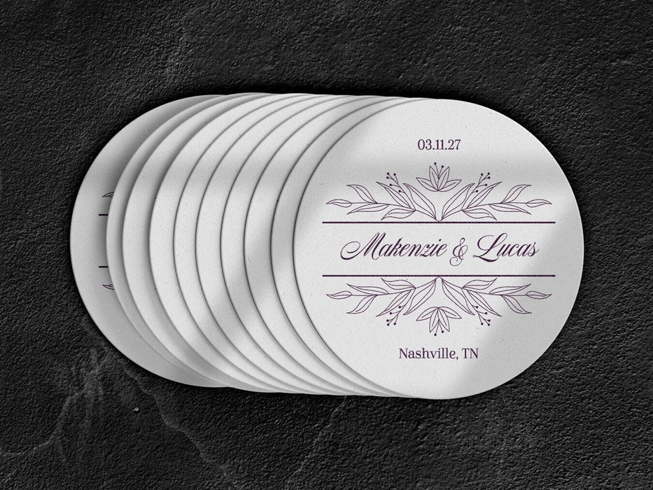 A stack of white coasters sitting on top of a black surface. Coasters feature a personalized floral design with the happy couple's first names, wedding date, and location.