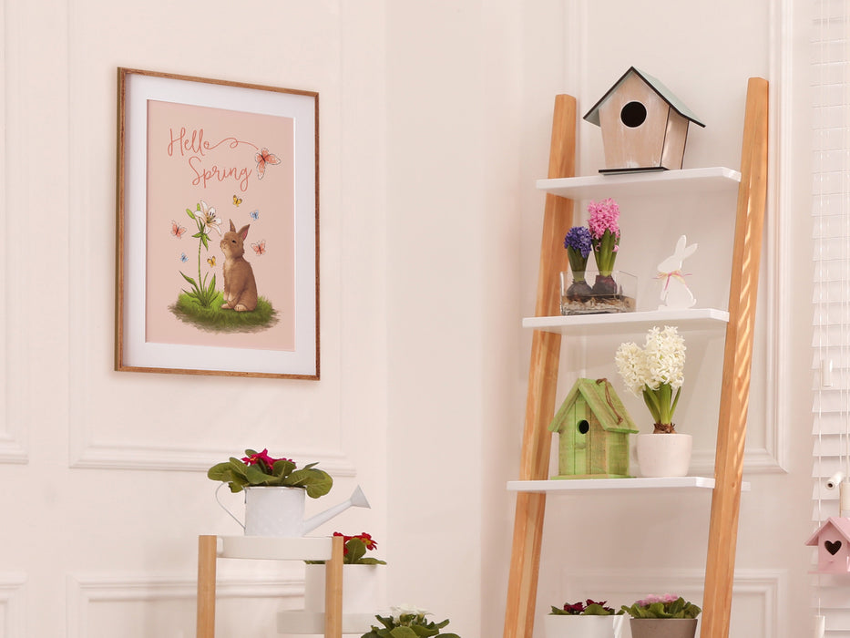 pastel easter print that says hello spring with a bunny and a lily surrounded by colorful butterflies in a wooden frame on white wall in living room surrounded by easter decor such as potted plants, flowers, and bird houses,