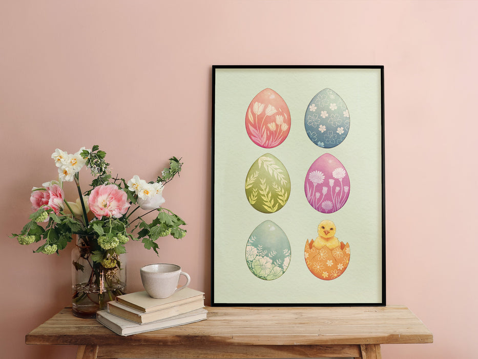 pastel easter art print of decorated eggs with a baby chick in black frame sitting ontop of wooden table next to potted flowers and a book and ceramic tea cup