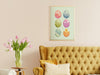pastel easter art print of decorated eggs with a baby chick in a gold frame on a white living room wall surrounded by decor and furniture such as a couch, pillows, a side table, and tulips in a vase
