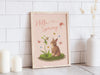 wooden frame with a pastel easter print that says hello spring with a bunny and a lily surrounded by colorful butterflies on a white kitchen counter top surrounded by candles and a potted plant