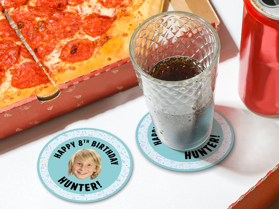 One coaster has a drink on it and an empty coaster sits beside it on a white table with pizza boxes. Coasters are designed with multi-colored confetti, teal blues, and a custom face photo. Coaster text reads Happy 8th Birthday Hunter!