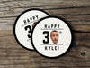 Two coasters sit on a wooden surface. Coasters are shown with custom face and text design. Coasters read Happy 30th Kyle! Coasters are designed with black text and a custom face photo.
