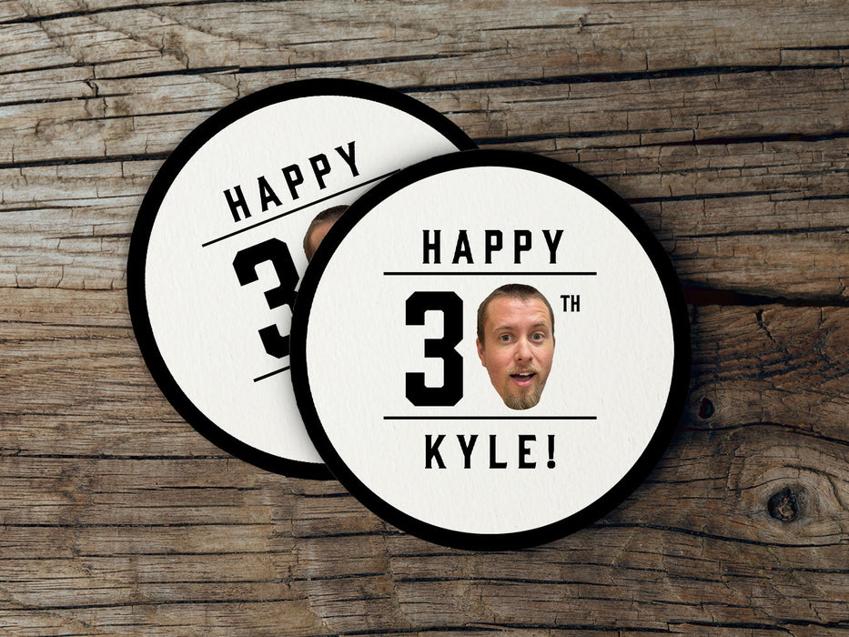 Two coasters sit on a wooden surface. Coasters are shown with custom face and text design. Coasters read Happy 30th Kyle! Coasters are designed with black text and a custom face photo.