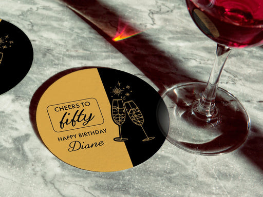 Two coasters are shown on a marble surface. One has a wine glass on it. Coasters are designed in black and gold ink with cheersing champagne glasses. Coaster text reads Cheers to Fifty. Happy Birthday Diane.
