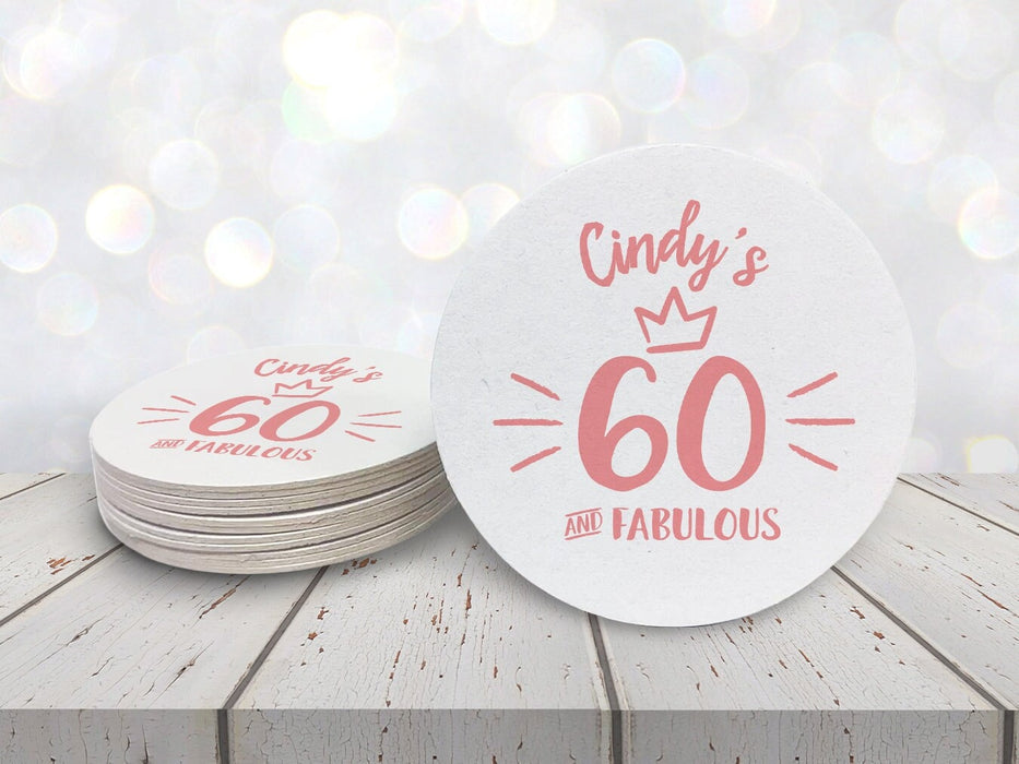 A stack of coasters by a single coaster sit on a white wooden table against a white glitter background. Coasters are designed with pink ink. Coaster text reads Cindy's 60 and Fabulous.