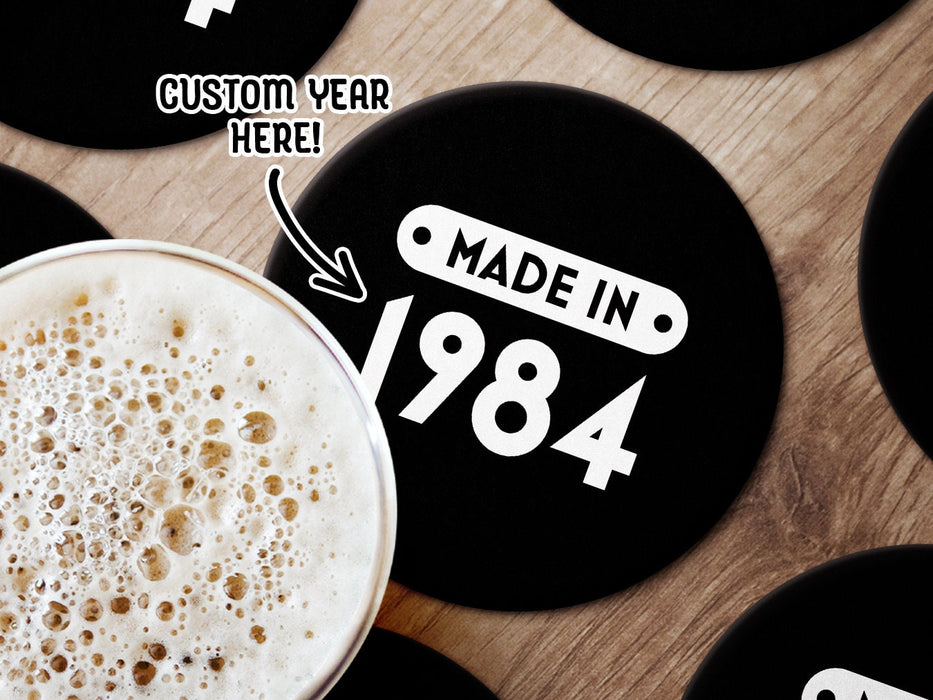 Coasters are on a wooden surface with a beer in the left bottom corner. Coasters are designed with a custom year. Coasters read MADE IN 1984 and designed with white text and black background.