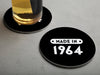One empty coaster is shown with a beer glass on a second coaster. Coasters are designed with a custom year. Coasters read MADE IN 1964 and designed with white text and black background.
