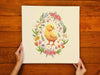 woman&#39;s hands holding a 12x12 inch canvas with spring easter chick pastel easter art on a brown wooden table
