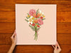 woman&#39;s hands holding a 12x12 inch canvas with spring bouquet pastel easter art on a brown wooden table