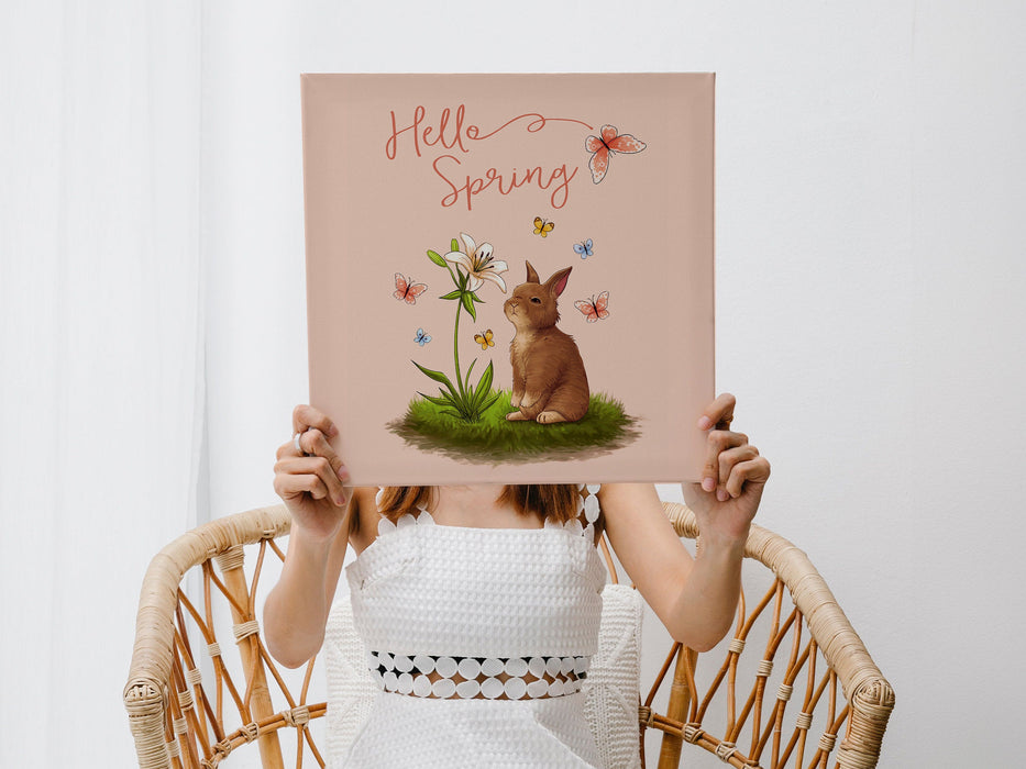 woman in white dress sitting in a wicker chair in a living room holding a 12x12 canvas with Hello Spring Easter print with a bunny and butterflies